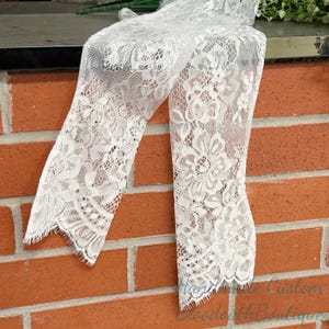 May include: Off-white lace fabric with a floral pattern. The delicate material is draped over a dark surface, with the edges of the fabric visible. The lace is against a red brick wall. The text "Handmade Custom Bloodtooth Boutique" is visible.
