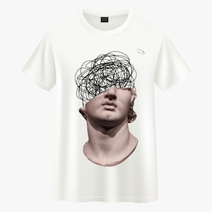 May include: White t-shirt with a graphic of a classical bust overlaid with a black scribble design. The bust is a pale pink, and the shirt has a crew neck and short sleeves. The design is a contemporary interpretation of classical art.