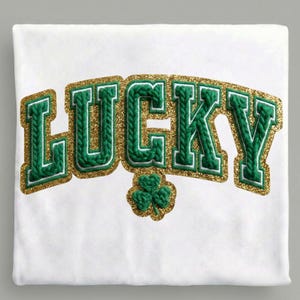 May include: White t-shirt with the word "LUCKY" in green, textured lettering outlined in gold glitter. A small green shamrock is below the text, also with gold glitter accents. The design is centered on the shirt.