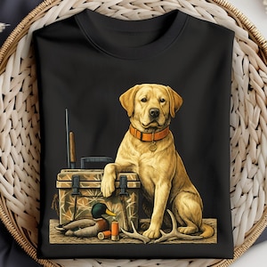 Op de afbeelding: Zwart sweatshirt met een gele Labrador Retriever met een oranje halsband. De hond zit naast een camouflage jachtkist, een geweer en een eendenlokker. Geweien en hagelpatronen zijn ook aanwezig.
