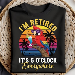 May include: Black t-shirt featuring a colorful parrot wearing sunglasses, holding a cocktail, and perched on a branch. The shirt has the text "I'm Retired, It's 5 O'Clock Everywhere" in yellow and white, with a sunset and palm tree background.