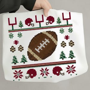 May include: White tote bag with a festive football-themed design. The bag features a brown football, red helmets, green Christmas trees, and goalposts. The design is reminiscent of a holiday sweater.