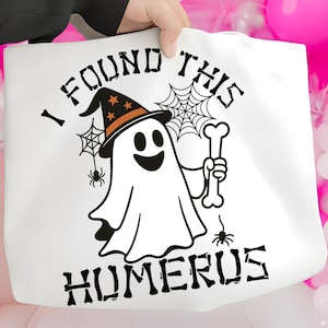 May include: White tote bag featuring a cartoon ghost wearing a witch hat and holding a bone. The text "I FOUND THIS HUMERUS" is printed in black. Spiders and spiderwebs are also included in the design.