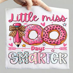 Puede incluir: Camiseta blanca con un diseño rosa. El diseño incluye el texto "Little Miss 100 Days Smarter" en rosa, junto con ilustraciones de donuts, un lápiz con un lazo de leopardo, corazones y estrellas.
