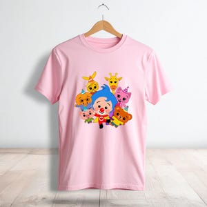 May include: A light pink t-shirt featuring a colorful graphic of cartoon characters. The design includes a clown and several animal figures. The shirt is hanging on a wooden hanger against a white wall.