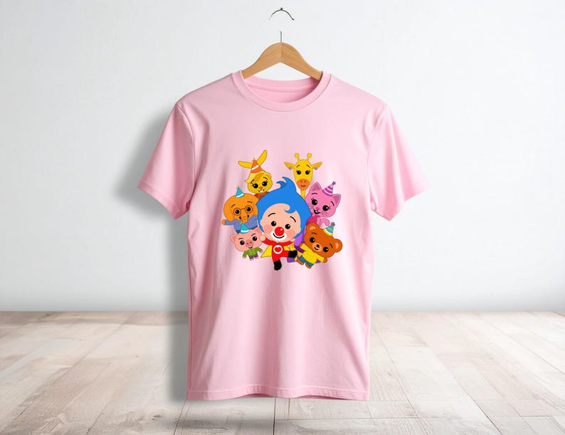 May include: A light pink t-shirt featuring a colorful graphic of cartoon characters. The design includes a clown and several animal figures. The shirt is hanging on a wooden hanger against a white wall.