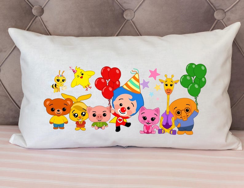 May include: White rectangular pillow with a colorful cartoon design featuring various animals and characters. The design includes a bee, a yellow bird, a bear, a pig, a clown, a cat, a giraffe, and an elephant, all holding balloons. The pillow is on a pink striped surface.