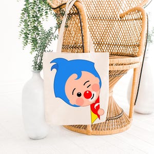 May include: A cream-colored tote bag featuring a cartoon character with blue hair, a red nose, and a red and yellow outfit. The bag has two handles and is displayed in front of a wicker chair and white vases with greenery.