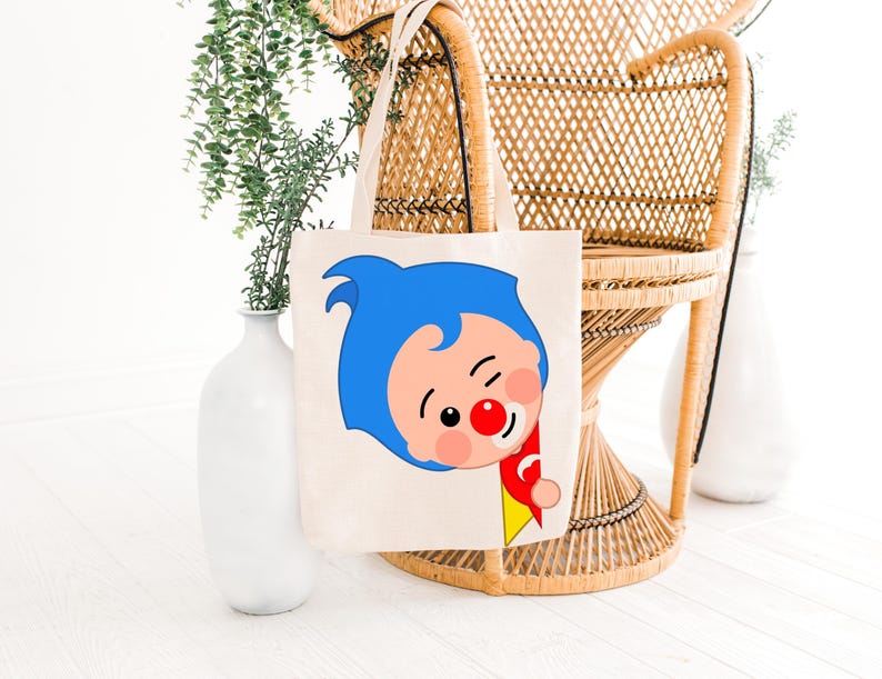 May include: A cream-colored tote bag featuring a cartoon character with blue hair, a red nose, and a red and yellow outfit. The bag has two handles and is displayed in front of a wicker chair and white vases with greenery.