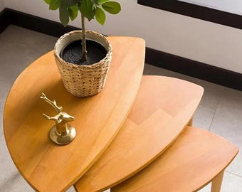 Pode incluir: Um conjunto de três mesas de encaixe de madeira com acabamento castanho claro. Uma pequena planta em vaso numa cesta de vime está sobre a mesa superior, e uma figura de veado dourada também é exibida. As mesas têm bordas arredondadas e pernas finas.