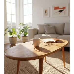 Puede incluir: Una mesa de centro ovalada de madera clara con cuatro patas cónicas. Una taza blanca, un libro abierto y gafas descansan sobre la mesa. La mesa está sobre una alfombra beige en una sala de estar luminosa con un sofá y obras de arte.