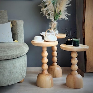 Puede incluir: Tres mesas auxiliares de madera clara con un diseño único. Cada mesa tiene una parte superior redonda y una base con una serie de elementos esféricos apilados. Una mesa tiene una tetera y una taza blancas. Las mesas están en una habitación con una pared gris y un sofá.