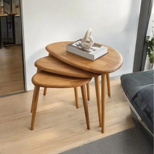Puede incluir: Un juego de tres mesas nido de madera con acabado marrón claro. Las mesas tienen una forma orgánica única y patas cónicas. Una escultura blanca decorativa y un libro están sobre la mesa superior. Las mesas están dispuestas sobre un suelo de madera clara.