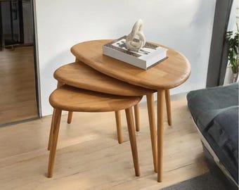 Pode incluir: Um conjunto de três mesas de encaixe de madeira com acabamento castanho claro. As mesas têm uma forma orgânica única e pernas afiladas. Uma escultura branca decorativa e um livro estão sobre a mesa superior. As mesas estão dispostas sobre um chão de madeira clara.