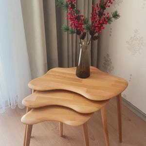 Puede incluir: Un conjunto de tres mesas nido de madera clara con una forma orgánica única. Las mesas tienen patas delgadas y cónicas. Un jarrón de cristal con bayas rojas y ramitas verdes se encuentra sobre la mesa más grande, añadiendo color.