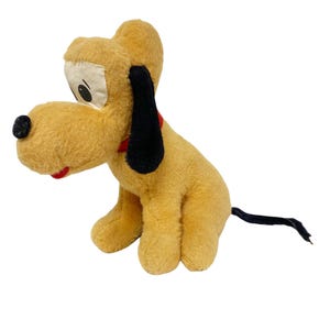 Pode incluir: Um peluche de cão com pelo amarelo dourado, apresentando um nariz preto, orelhas pretas compridas e uma cauda preta fina. A personagem tem uma mancha facial cor de creme com olhos pretos e uma coleira vermelha à volta do pescoço, sentado.