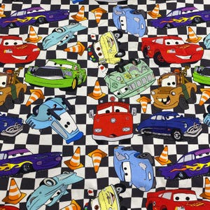 Puede incluir: Tela con un patrón repetido de coches de dibujos animados en varios colores, incluyendo rojo, azul, verde y morado. Los coches están sobre un fondo a cuadros blanco y negro, con conos de tráfico naranjas esparcidos.