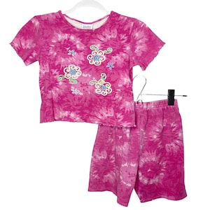 May include: A two-piece set of tie-dye clothing in shades of pink. The top is a short-sleeved shirt with embroidered floral designs. The matching shorts have an elastic waistband. The set is displayed on a white hanger.