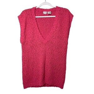 May include: A sleeveless, hot pink sweater with a deep V-neck. The sweater is made of a textured knit fabric and hangs on a white hanger. The garment has a tag at the neckline.