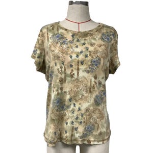 Puede incluir: Una camiseta de manga corta y cuello redondo con un estampado floral de cachemira en beige, marrón y azul. La camiseta se muestra en un maniquí. La tela tiene un aspecto suave y ligeramente descolorido.