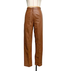 Könnte beinhalten: Braune Lederhose mit hohem Bund und geradem Bein. Die Hose hat einen Knopfverschluss und wird auf einer Schaufensterpuppe präsentiert. Das Leder hat eine mittelbraune Farbe.