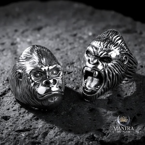 May include: Two silver gorilla head rings. One ring depicts a gorilla with a closed mouth, the other with an open mouth and visible teeth. The rings are on a dark, textured surface. The rings are likely made of stainless steel.