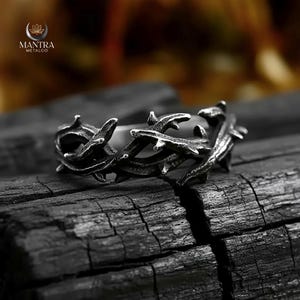 May include: A silver-toned ring with a thorny vine design. The ring is set against a dark, textured background. The ring's design features multiple thorn-like projections, creating a rugged, nature-inspired aesthetic. The brand logo "MANTRA METALCO" is visible in the upper left corner.