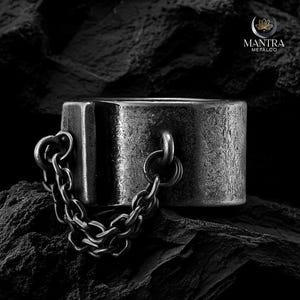 May include: A silver-toned ring with a chain attached. The ring is a thick, rectangular shape with a textured surface. The chain is made of small, interlocking links. The Mantra Metalco logo is in the upper right corner.