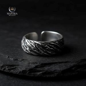 Op de afbeelding: Een zilverkleurige verstelbare ring met een textuur, organisch ontwerp. De ring heeft een gedetailleerd patroon dat om de band loopt. Het Mantra Metalco-logo is zichtbaar in de linkerbovenhoek.