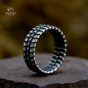 Peut inclure: Bague argentée avec un motif en écailles. Le design de la bague enveloppe tout le tour. La bague en métal est présentée sur une surface sombre et texturée. Le design de la bague rappelle les écailles de dragon.