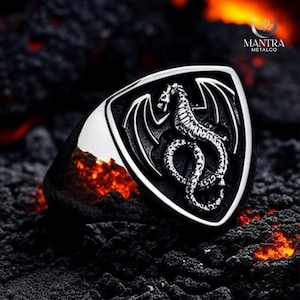 May include: A silver-colored ring with a shield-shaped face featuring a detailed dragon design. The dragon has wings and a serpentine body. The ring is set against a dark, textured background with hints of orange.