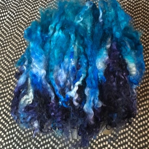 May include: A pile of blue, purple, and white curly yarn. The yarn is arranged in a loose, fluffy mound.