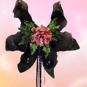 Puede incluir: Decoración en forma de mariposa negra con una flor rosa y hojas verdes en el centro. La mariposa está hecha de tela de malla negra con detalles de purpurina.