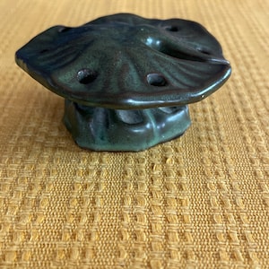 May include: A small, green ceramic mushroom-shaped trinket with a flat top and five holes. The mushroom is sitting on a yellow woven surface.