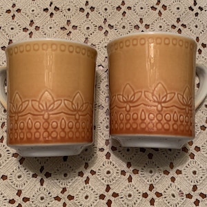 May include: Two brown ceramic mugs with a decorative pattern around the top and bottom. The mugs have a white interior and a white base.