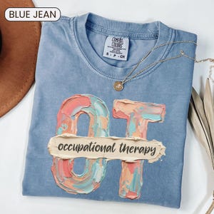 May include: A blue jean colored t-shirt with a crew neckline and a Comfort Colors label. The graphic features the letters OT in a textured, multi-colored paint stroke style, overlaid with a horizontal beige brushstroke reading occupational therapy.