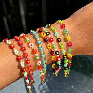 Könnte beinhalten: Eine Sammlung von Armbändern mit Perlen, die jeweils mit farbenfrohen Blumenmotiven verziert sind. Die Armbänder weisen rote, weiße, blaue, gelbe und grüne Blumenmuster auf. Goldfarbene Verschlüsse hängen von den Armbändern herab und verleihen ihnen einen Hauch von Eleganz.