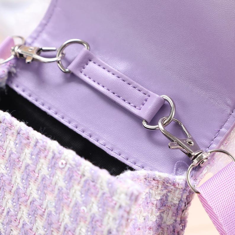 K&ouml;nnte beinhalten: Eine lavendelfarbene Umh&auml;ngetasche aus Tweed mit einem strukturierten Webmuster und dezenten Pailletten. Die Tasche hat eine glatte, lavendelfarbene Innenklappe, ein schwarzes Innenfutter und silberfarbene Karabinerhaken aus Metall.
