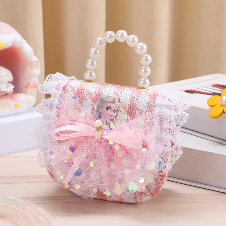 K&ouml;nnte beinhalten: Eine kleine Handtasche mit rosa-wei&szlig;em Tweedmuster, wei&szlig;er Spitzenborte und einem Perlenhenkel. Die Vorderseite ist mit einer rosa T&uuml;llschleife, irisierenden Pailletten und einer Grafik einer blonden Figur in einem blauen Kleid verziert.