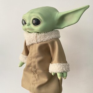 May include: A green creature figurine with large black eyes and pointed ears, wearing a tan coat with a white textured sherpa collar and matching sleeve cuffs. The figure has a smooth skin texture and is shown from a side profile angle.