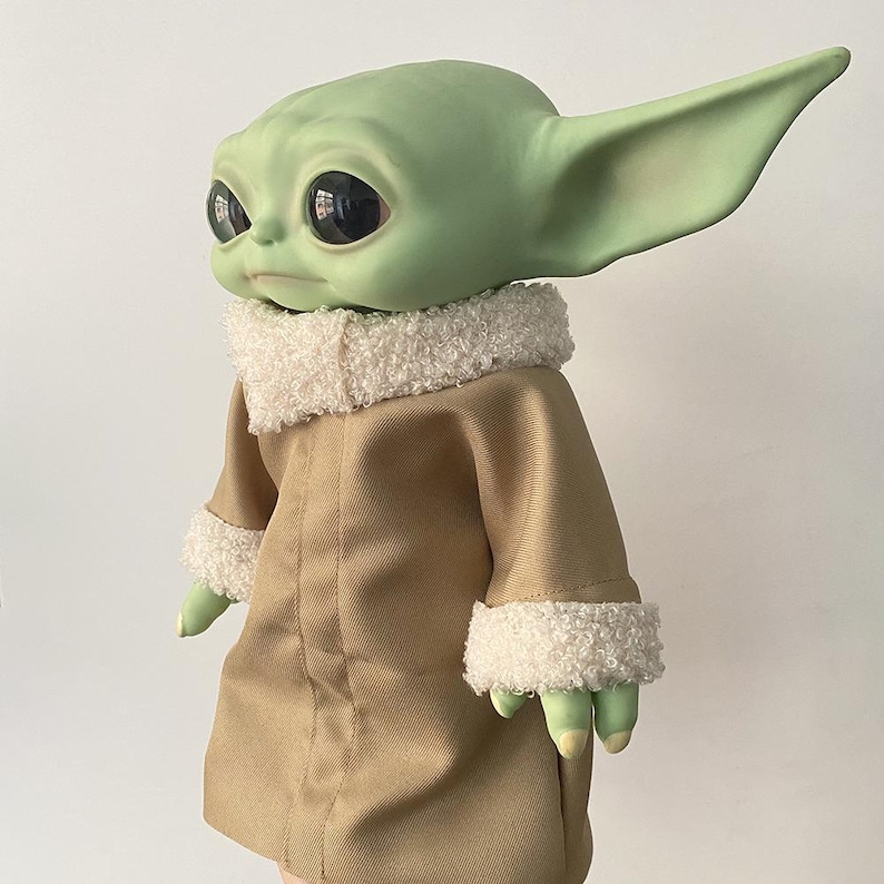 May include: A green creature figurine with large black eyes and pointed ears, wearing a tan coat with a white textured sherpa collar and matching sleeve cuffs. The figure has a smooth skin texture and is shown from a side profile angle.