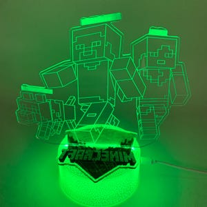 以下が含まれることがあります： 緑色に光るMinecraftをテーマにしたナイトライト。ピクセル化されたキャラクターとMinecraftのロゴが特徴です。ライトのベースはテクスチャ加工された球体で、キャラクターは明るい緑色で縁取られています。