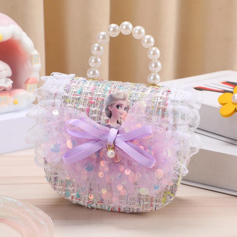 K&ouml;nnte beinhalten: Eine kleine Handtasche aus mehrfarbigem Tweed-Stoff mit einem wei&szlig;en Perlenhenkel und wei&szlig;er Spitzenborte. Die Vorderseite ist mit einer blonden Figur, einer lavendelfarbenen Schleife und einer Lage aus rosa T&uuml;ll mit irisierenden Pailletten verziert.