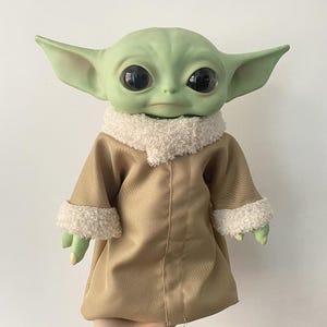 May include: A green creature doll with large black eyes and pointed ears, wearing a tan coat with a white textured sherpa collar and matching white sherpa cuffs on the sleeves. The doll has a smooth skin texture and a neutral facial expression.