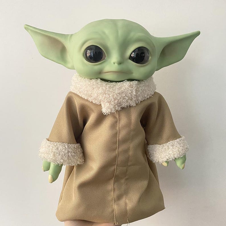 May include: A green creature doll with large black eyes and pointed ears, wearing a tan coat with a white textured sherpa collar and matching white sherpa cuffs on the sleeves. The doll has a smooth skin texture and a neutral facial expression.