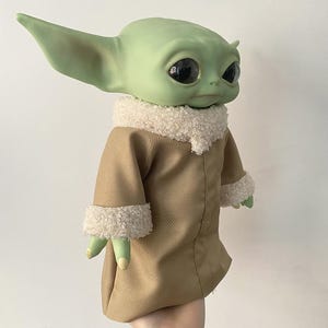 May include: A green creature figurine with large black eyes and pointed ears, wearing a tan coat with a white textured sherpa collar and matching cuffs. The figure has a smooth skin texture and is depicted from the chest up in a side profile.