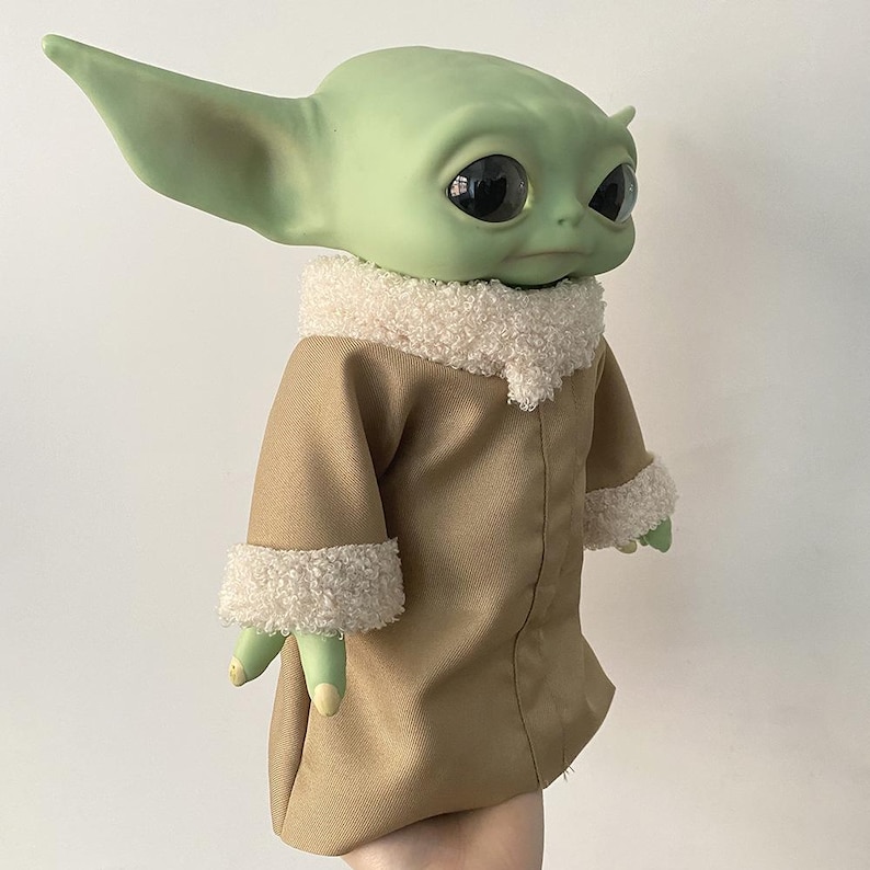 May include: A green creature figurine with large black eyes and pointed ears, wearing a tan coat with a white textured sherpa collar and matching cuffs. The figure has a smooth skin texture and is depicted from the chest up in a side profile.