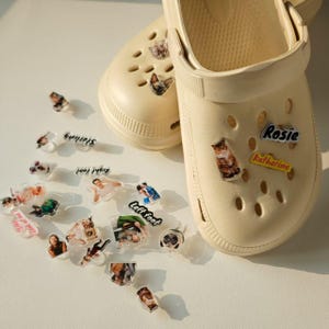 Puede incluir: Zuecos color crema con varios dijes decorativos esparcidos. Los dijes presentan imágenes de mascotas, personas y texto, incluyendo los nombres "Rosie" y "Katherine", y las palabras "Left foot" y "Right foot".