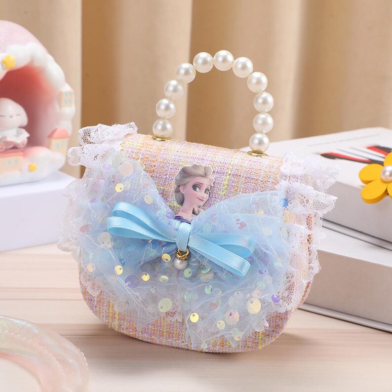 K&ouml;nnte beinhalten: Eine kleine Handtasche aus hellrosa und gelb kariertem Stoff mit einem wei&szlig;en Perlenhenkel und einer gro&szlig;en hellblauen T&uuml;llschleife mit irisierenden Pailletten. In der Mitte der Schleife befindet sich eine kleine Figur mit blondem Haar.