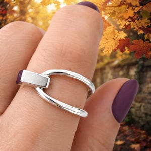 Puede incluir: Un anillo de metal plateado con un diseño liso y abierto, que presenta un elemento de barra rectangular que conecta ambos lados. El anillo tiene una superficie pulida y reflectante, con una banda curva continua que rodea el dedo.